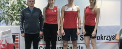 Cztery osoby w strojach sportowych stoją przed tablicą. Kluczowe obiekty: mężczyzna, trzy kobiety, stroje sportowe, tablica.