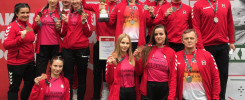 Grupa sportowców w czerwonych strojach odbiera medale i puchar. Na podium stoją młodzi ludzie i starszy mężczyzna.