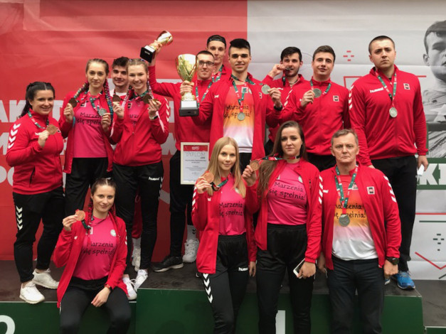 Grupa sportowców w czerwonych strojach odbiera medale i puchar. Na podium stoją młodzi ludzie i starszy mężczyzna.