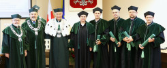 Grupa osób w togach akademickich stoi przed godłem Polski. Kluczowe obiekty: ludzie, togi, godło.