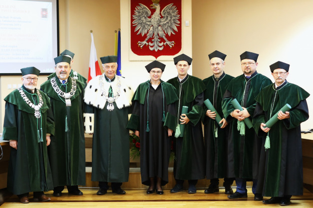 Grupa osób w togach akademickich stoi przed godłem Polski. Kluczowe obiekty: ludzie, togi, godło.