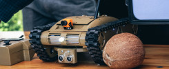 Gąsienicowy robot z kamerą i pilotem, obok orzecha kokosowego. Kontekst: wystawa, prezentacja technologii.