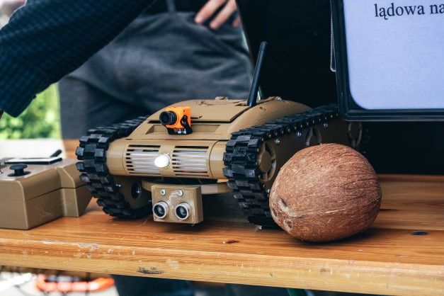 Gąsienicowy robot z kamerą i pilotem, obok orzecha kokosowego. Kontekst: wystawa, prezentacja technologii.
