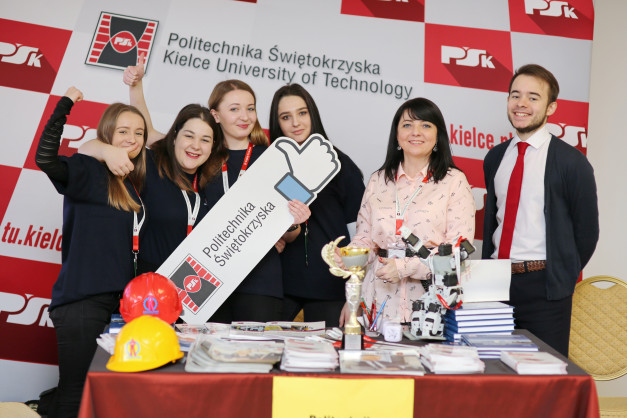 Grupa osób pozuje z tabliczką "Politechnika Świętokrzyska". W tle logo uczelni. Kluczowe obiekty: ludzie, tabliczka, logo.