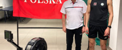 Dwaj mężczyźni stoją obok wioślarza. Na ścianie wisi biało-czerwona flaga z napisem "POLSKA".