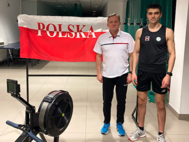 Dwaj mężczyźni stoją obok wioślarza. Na ścianie wisi biało-czerwona flaga z napisem "POLSKA".