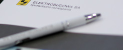 Biały długopis leży na papierze z logo ELEKTROBUDOWA SA i napisem "Sprawdzone rozwiązania".