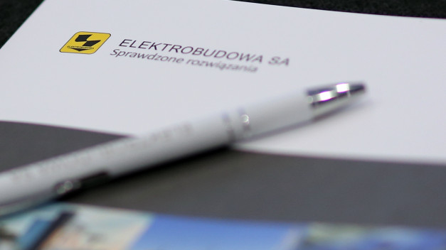 Biały długopis leży na papierze z logo ELEKTROBUDOWA SA i napisem "Sprawdzone rozwiązania".