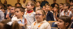 Dzieci w sali siedzą na krzesłach, niektóre podnoszą ręce. W tle widać dorosłych. Kontekst: wydarzenie edukacyjne.