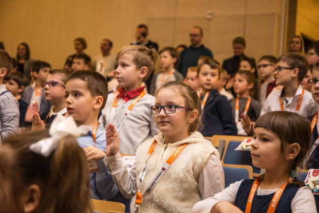Dzieci w sali siedzą na krzesłach, niektóre podnoszą ręce. W tle widać dorosłych. Kontekst: wydarzenie edukacyjne.