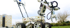 Robot z ramieniem i kołami na trawie, z budynkiem w tle. Kluczowe obiekty: robot, ramię, koła, budynek.
