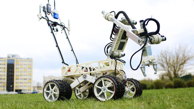 Robot z ramieniem i kołami na trawie, z budynkiem w tle. Kluczowe obiekty: robot, ramię, koła, budynek.