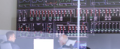 Duży ekran wyświetla schematy elektryczne i wykresy. Obraz zawiera ludzi obserwujących monitor.
