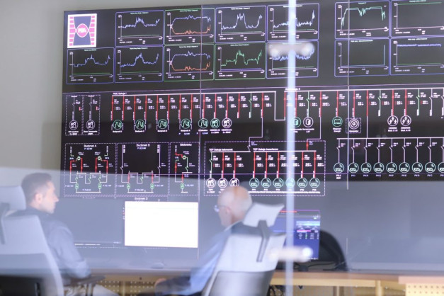 Duży ekran wyświetla schematy elektryczne i wykresy. Obraz zawiera ludzi obserwujących monitor.