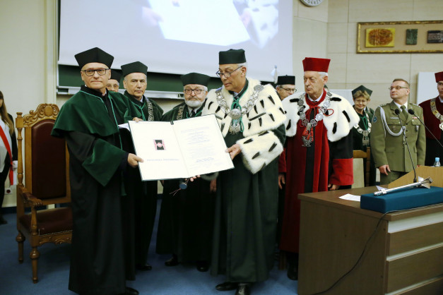 Uroczystość wręczenia dyplomów. Kluczowe obiekty: dyplom, togi, czapki, łańcuchy. Kontekst: akademicki.