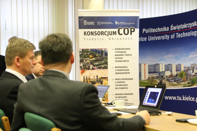 Mężczyźni w garniturach siedzą przy stole, przed nimi baner z logo uczelni i napisem "KONSORCJUM COP".