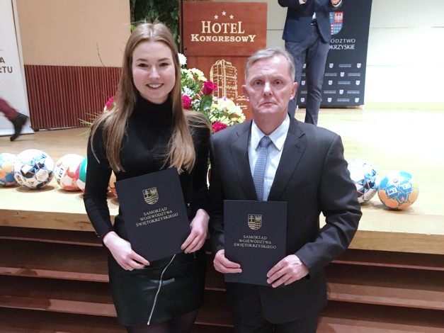 Kobieta i mężczyzna trzymają dokumenty. W tle widoczne piłki, logo hotelu i baner.