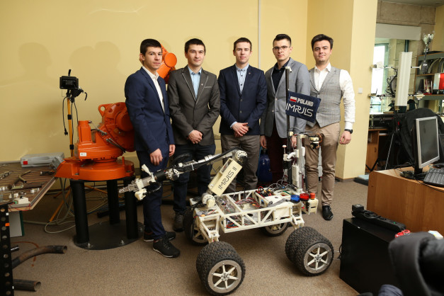 Pięciu mężczyzn stoi obok robota mobilnego i ramienia robota. Kontekst to prawdopodobnie projekt inżynieryjny lub edukacyjny.