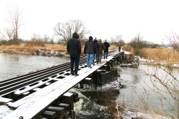 Grupa ludzi idzie po zasypanym śniegiem moście kolejowym nad rzeką.