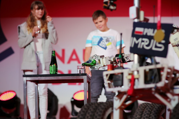 Robot nalewa płyn z butelki do szklanki. W tle dziewczyna i chłopiec.