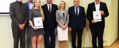 Grupa osób w strojach formalnych stoi na tle ściany. W rękach trzymają dokumenty.