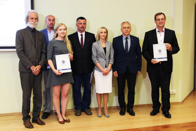 Grupa osób w strojach formalnych stoi na tle ściany. W rękach trzymają dokumenty.