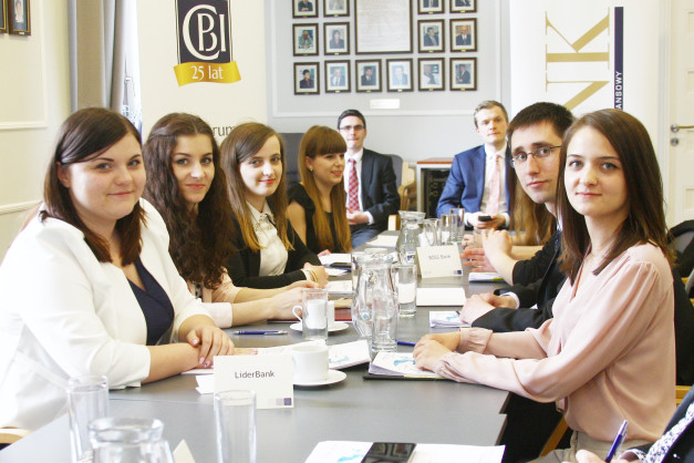 Osoby siedzą przy stole konferencyjnym. Kluczowe obiekty: ludzie, stoły, dokumenty, logo "LiderBank".