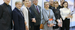 Grupa osób pozuje do zdjęcia. Widać mężczyzn i kobiety w eleganckich strojach, stojących przed tablicami informacyjnymi.