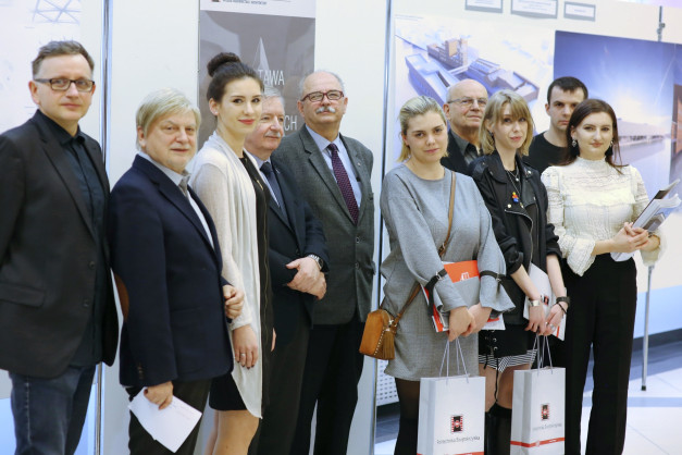 Grupa osób pozuje do zdjęcia. Widać mężczyzn i kobiety w eleganckich strojach, stojących przed tablicami informacyjnymi.