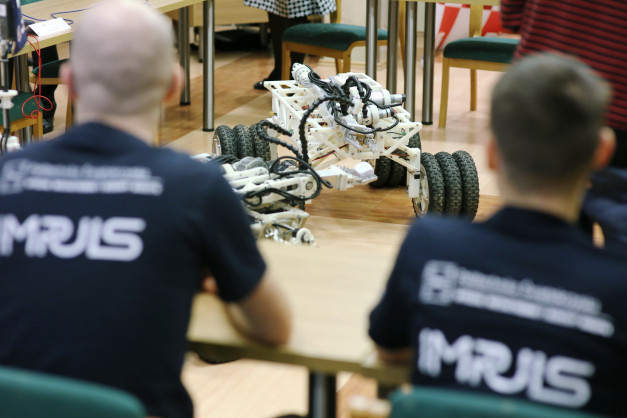 Dwaj mężczyźni w koszulkach z napisem "MRJS" obserwują robota. Kontekst: zawody robotyczne.