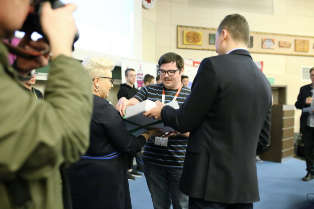 Ludzie odbierają i wręczają pudełko. W tle widać obrazy i uczestników.