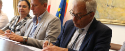 Trzech ludzi przy stole podpisuje dokumenty. W tle flagi Polski i UE, obraz.