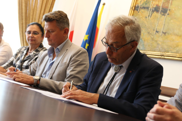 Trzech ludzi przy stole podpisuje dokumenty. W tle flagi Polski i UE, obraz.