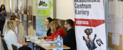 Ludzie siedzą przy stołach, obok banerów informacyjnych. Kontekst: targi pracy lub wydarzenie edukacyjne.
