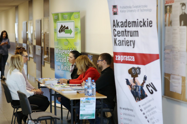 Ludzie siedzą przy stołach, obok banerów informacyjnych. Kontekst: targi pracy lub wydarzenie edukacyjne.
