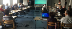Ludzie siedzą przy stołach podczas prezentacji. Widać ekran projekcyjny, flipchart i laptopy.