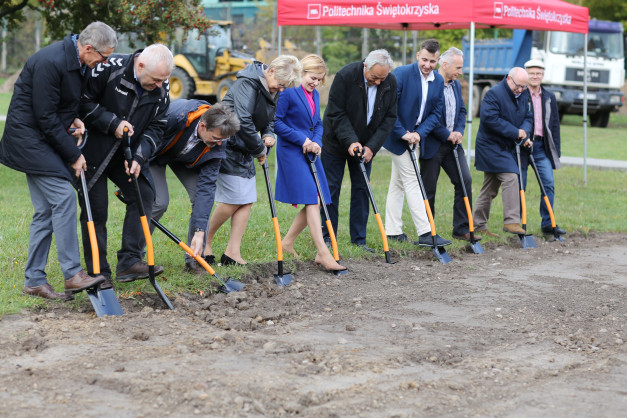 Grupa osób w strojach formalnych i casualowych trzyma łopaty, gotowa do symbolicznego wbicia ich w ziemię.