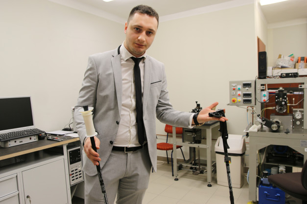Mężczyzna w garniturze trzyma kijki trekkingowe. W tle widać sprzęt laboratoryjny i komputer.