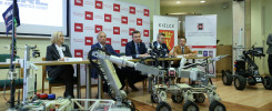 Konferencja prasowa z robotami mobilnymi. Na stole siedzą ludzie, przed nimi roboty.