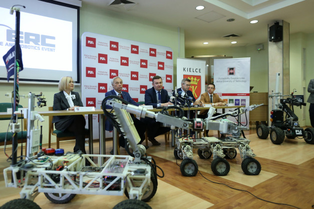 Konferencja prasowa z robotami mobilnymi. Na stole siedzą ludzie, przed nimi roboty.