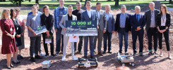 Grupa osób pozuje do zdjęcia z nagrodami i robotami. Kluczowe obiekty: ludzie, tablice z nagrodami, roboty.