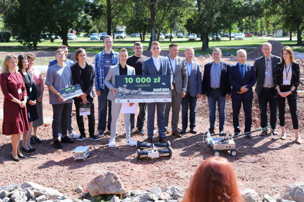 Grupa osób pozuje do zdjęcia z nagrodami i robotami. Kluczowe obiekty: ludzie, tablice z nagrodami, roboty.