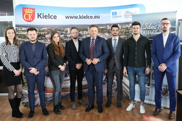 Grupa osób pozuje przed banerem z logo Kielc. W tle widoczne miasto.