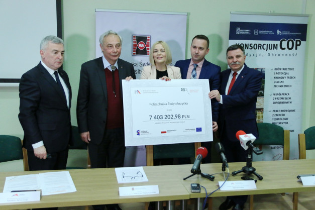 Cztery osoby stoją za stołem, trzymając czek na kwotę 7 403 202,98 PLN. Kontekst to wręczenie grantu.