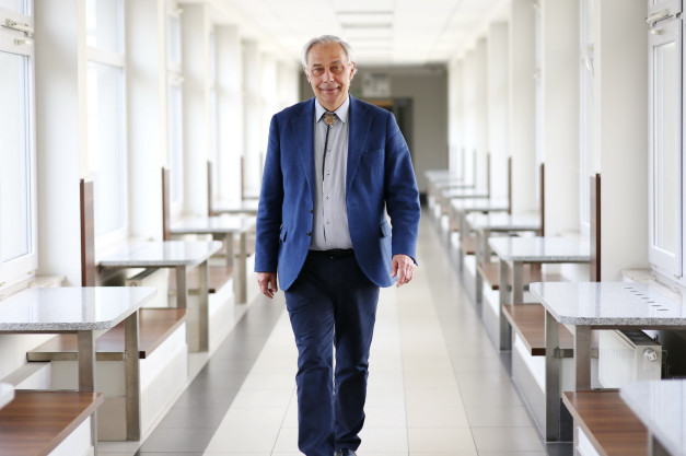 Starszy mężczyzna w niebieskiej marynarce idzie korytarzem z rzędami stołów i ławek.