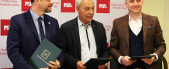 Trzech mężczyzn w garniturach stoi przed ścianą z logo Politechniki Świętokrzyskiej. Trzymają zielone teczki.