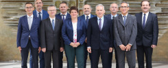 Grupa dziesięciu osób w formalnych strojach stoi przed kamienną ścianą. Wszyscy ubrani są w garnitury i garsonki.