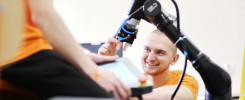 Mężczyzna w pomarańczowej koszulce pracuje przy ramieniu robota. W tle widać tablet.