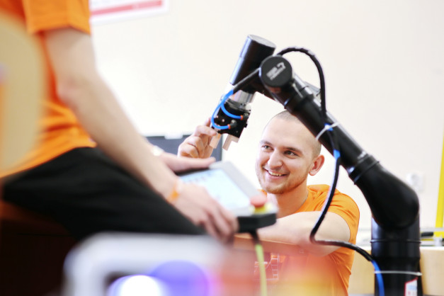 Mężczyzna w pomarańczowej koszulce pracuje przy ramieniu robota. W tle widać tablet.
