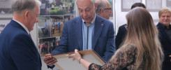 Dwóch mężczyzn i kobieta w galerii sztuki. Mężczyzna w garniturze wręcza ramę z obrazem.